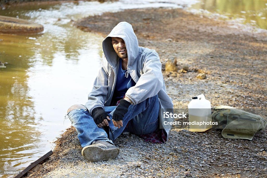 Homeless man Young homeless man sitting on river bank in damaged area Abandoned Stock Photo