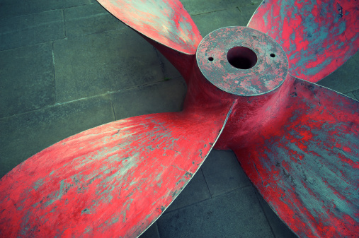 Massive propeller in distressed red paint sits on stone background
