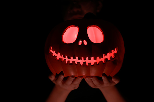 A lantern carved from pumpkin known as a Jack-o-lantern glows in hand with  the dark on a black background