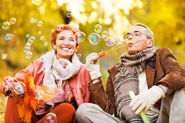alegre pareja senior soplando burbujas en el parque - autumn women leaf scarf fotografías e imágenes de stock