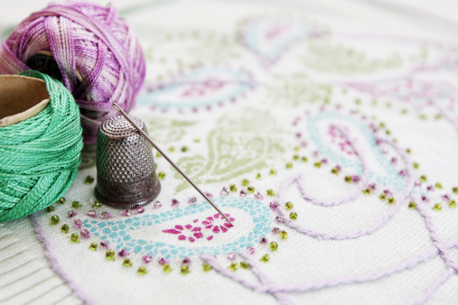 A studio shot of a embroidery with two embroidery thread standing on the left featuring green and purple colours with a needle and thimble on front.