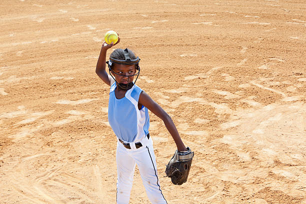 Softball player throwing ball African American girl (9 years), infield player, throwing softball.  Wearing face mask, standard safety equipment required by some leagues. softball pitcher stock pictures, royalty-free photos & images