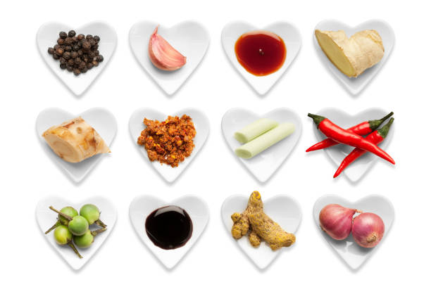 Traditional Thai Ingredients in heart shaped bowls isolated on a white background with a clipping path. . In order from top left corner horizontally - Whole black peppercorns, Garlic Clove, Light Soya Sauce, Fresh Ginger, Galangal Root, Chilli Paste, Lemongrass, Fresh Chillies, Pea Aubergine, Dark Soya Sauce, Turmeric, Shallots.