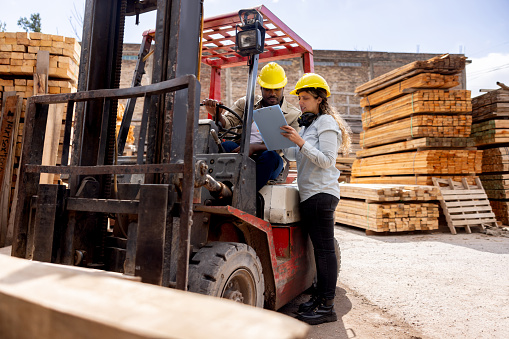 Latin American female foreperson talking to the forklift driver at a lumberyard and coordinating the transportation of wood