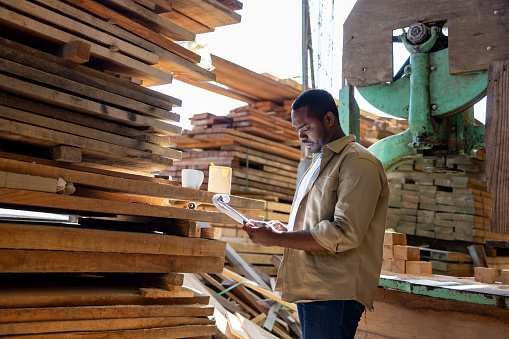 African American man working at a lumberyard taking inventory of the wood and writing on a clipboard - people at work concepts