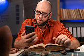 Archeologist working late night in office uses smartphone and old science book