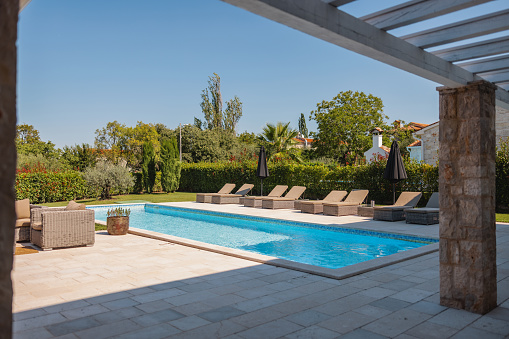 A beautiful backyard of a vacation rental with a large pool and a charming terrace. There are brown chairs by the pool as well as a seating area. The sun is shining and the sky is blue. The light is illuminating the pool and its surroundings.