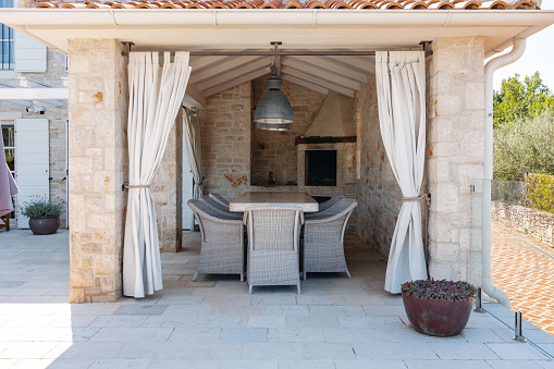 Covered seating area in the backyard of a beautiful Mediterranean-style villa. The exterior of the building is made out of stone. The chairs are made of ratan and the table is made of stone. There are two pendant lights over the big table.