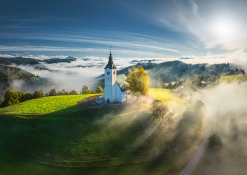 Aerial view of small church on the hill in low clouds at sunrise in autumn. Slovenia. Top view of beautiful chapel on mountain in fog, green alpine meadows, trees, blue sky at dawn in fall. Travel