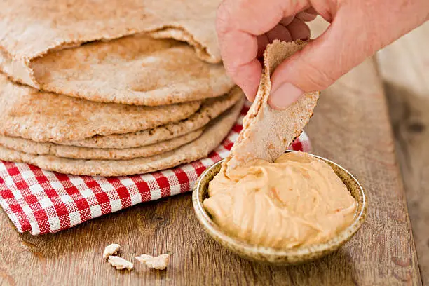 A close up shot of a hand dipping a piece of pitta brad into some hummus. Shot against a wooden background.