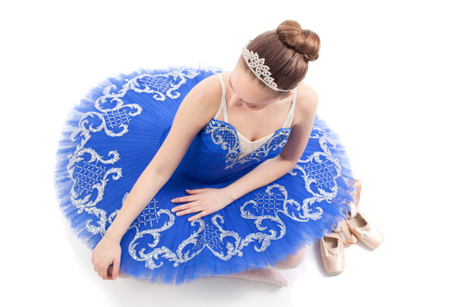 Classic ballerina sitting on floor in her blue tutu dress on white background. The danceraas cream satin ballet Pointe shoes are beside her. Look from above.