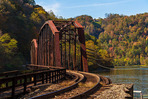 A rusted railroad trestle over a river in West Virginia.  The train tracks that go through the trestle are still in use.  Sun was setting over the mountain to the left and hazy fall colors can be seen on trees near the water.