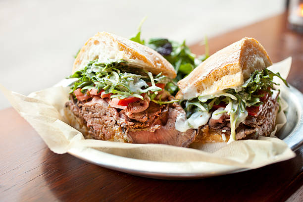 Roast beef sandwich "Freshly roasted beef sliced very thin and piled high onto a sweet baguette; served with organic greens, caramelized onions, peppers and horseradish aioli." delicatessen stock pictures, royalty-free photos & images