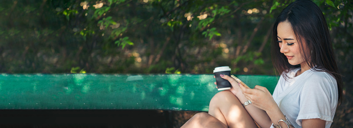 Banner Woman use smartphone drinking coffee hand holding hot disposable cup in green park. Web Banner Happy Relax asian woman smiling face at outdoors garden. Young women enjoy nature with copy space