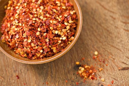 A high angle close up of a small brown bowl full of crushed red pepper flakes. Shot on a wooden background.