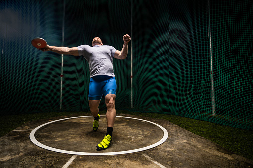 Muscular male athlete throwing discus, practicing for track and field event at night