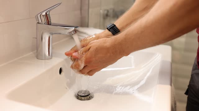 Young man puts hands under running tap water and cleanses skin in bathroom and does morning routine