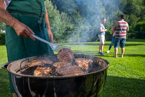 Photo of a man grilling meat on an outdoor coal barbecue with people talking on the lawn in the background.