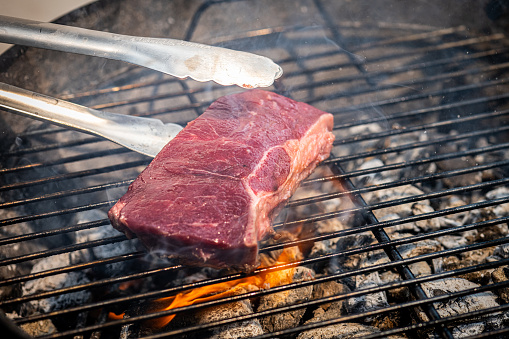 Close up photo of tongs grabbing a beef steak roasting on a barbecue charcoal grill.