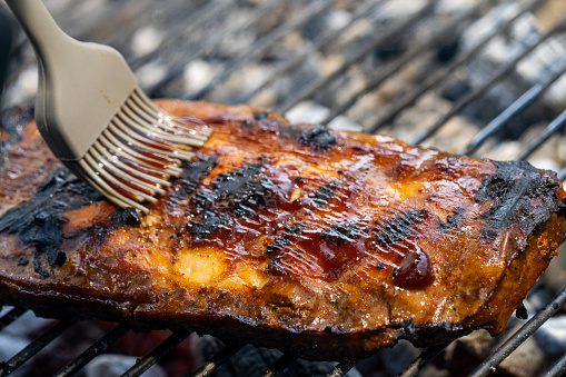 Pork ribs on a charcoal barbecue grill getting smeared with gravy using a grill brush.