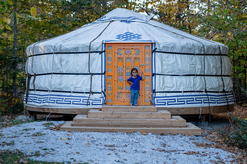 A sweet little girl poses at the door of her families yurt during a camping trip.  She is dressed casually and the autumn leaves can be seen setting in.
