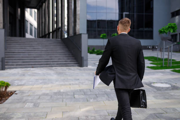 istock Full rear view of businessman with briefcase and folder with documents in hands walking towards office building 1755204584