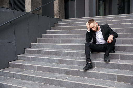 The dismissed businessman sits upset on the stairs near the office building. He lost his job. The unemployment rate is on the rise due to the pandemic.