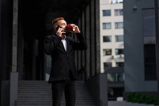 A young business man holds coffee in his hand and speaks on a cell phone.