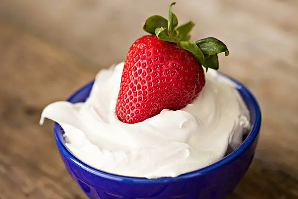 A close up shot of a red strawberry dipped in white whipping cream in a small blue bowl. The perfect dessert for an American patriotic holiday.