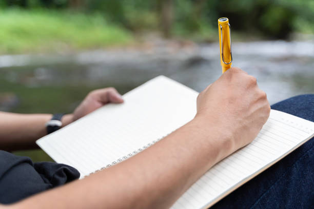 istock Young man takes notes in a notebook on nature. Man sits by a tree with his back to him, a pencil and a notebook in his hands. 1755237703