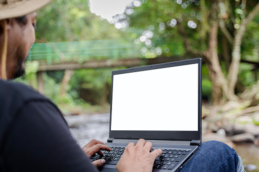 Young man using a laptop computer Notebook outside in the middle of nature against the background of the mountain forest. Online distance work. Copy space for design or text on white laptop screen.