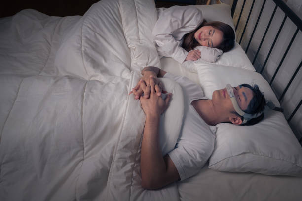 Happy Asian couple with CPAP machine on side table with husband wearing CPAP mask sleeping smoothly in bed all night without snoring stock photo