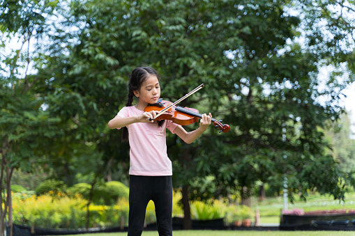 A cute white Asian girl is playing the violin in the middle of beautiful nature, greenery and sunlight. She is learning about new things that interest her.