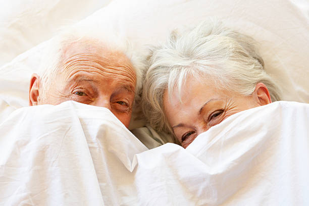 Senior Couple Relaxing In Bed Hiding Under Sheets Senior Couple Relaxing In Bed Hiding Under Sheets Smiling And Laughing. old man pajamas photos stock pictures, royalty-free photos & images