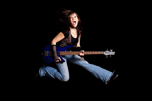 Color image of a tough rocker girl holding a guitar and jumping up in the air, with black background.