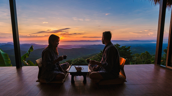 a couple of men and women on vacation in Northern Thailand , staying at a homestay cabin hut in the mountains of Chiang Rai Doi Chang drinking tea on the wooden balcony at sunrise