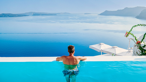 a young men in swim shorts relaxing in the pool looking out over the caldera of Santorini Island Greece, man at an infinity pool, a young guy on a luxury vacation in Europe Greece.