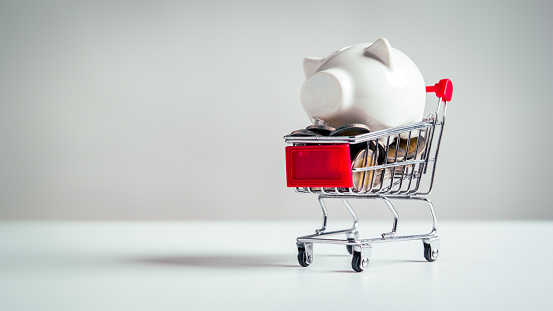 Piggy bank in shopping cart with plenty full of coins on white table background. Pig jar money savings and wealth accumulation collecting concept.