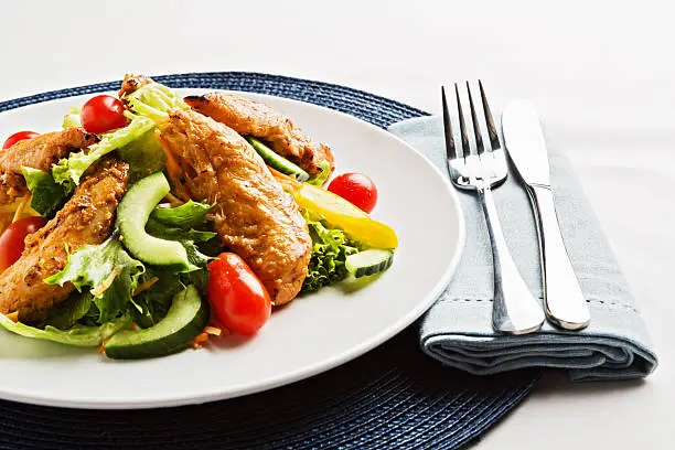 "Tandoori chicken salad, with cherry tomatoes, cucumber, sweet yellow bell peppers and lettuce, sitting on a gray grass tablemat with a linen napkin and stainless steel silverware."