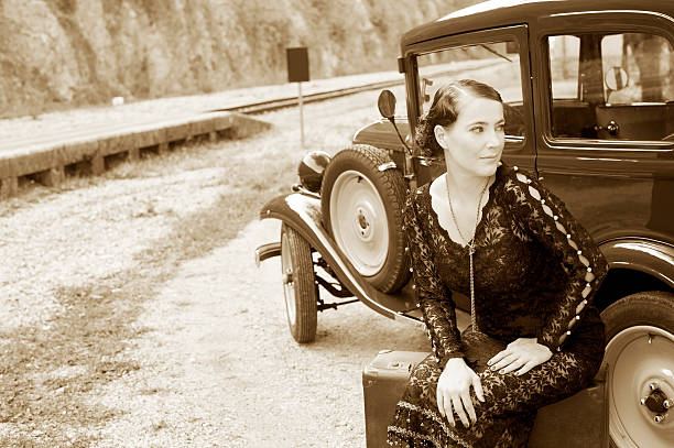 Woman sitting in front a vintage car on abig suitcase and looking around. 1939 style. Sepia toned.