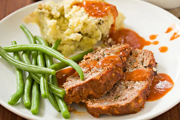 "A close up shot of a meatloaf dinner, two slices of meatloaf, skin on mashed potatoes, lightly steamed green beans and a tomato sauce."