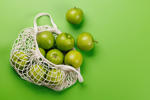 Mesh bag with fresh green apples over green background. Flat lay with copy space