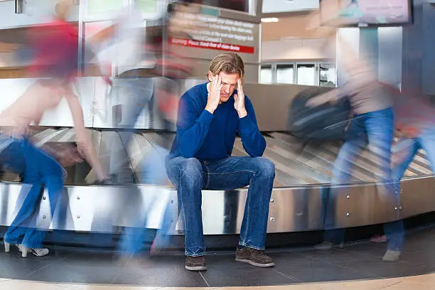 Lost baggage in a busy airport. Man is sitting on a baggage claim carousel rubbing his head. He has a head ache. Nearby people picking up their luggage are depicted by motion blur movement. Traveler is frustrated and sad.  