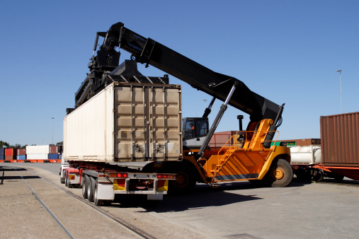 Transferring freight containers from train to truck at a city intermodal hub.  A modern container stacker loads a freight container onto a truck on a hardstand container terminal.  The container has come from a train to the left of the frame.  In the background are rail wagons (freight cars) loaded with a variety of containers. Logos and trademarks have been removed.