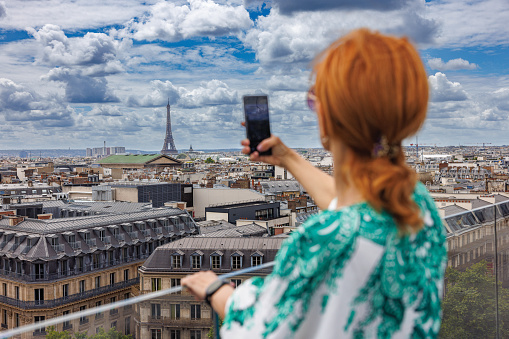 Rear view on beautiful woman standing on the high terrace and taking pictures of cityscape with her smart phone