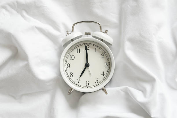 istock White alarm clock in bed with a white blanket close-up. 1755522711