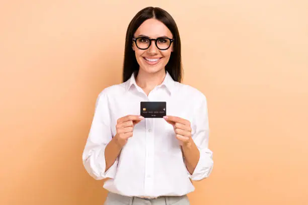 Photo of cheerful smiling lady wear white shirt spectacles showing holding bank card isolated beige color background.