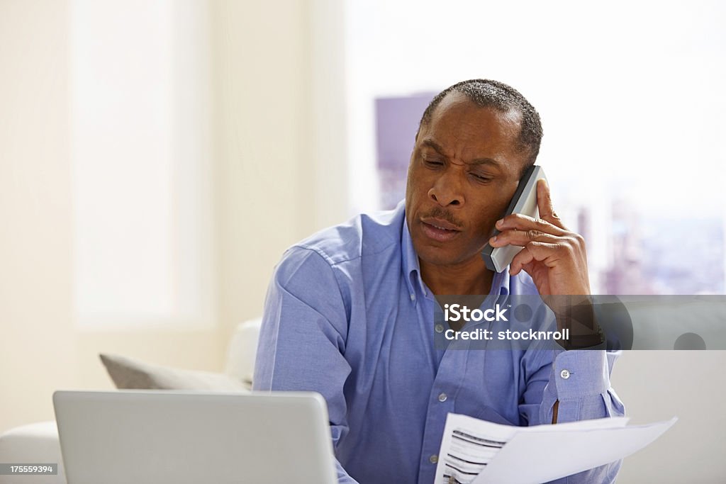 African man calculating his monthly expenses Image of mature African man holding papers and talking on phone while sitting at home with a laptop African-American Ethnicity Stock Photo