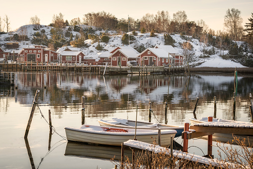Small rowboats moored at a jetty. Idyllic cottages on small island in the back. Snow on the ground in Swedish winter.