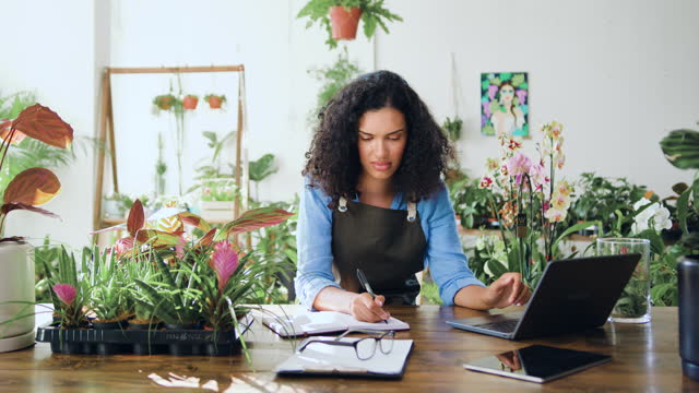 African american woman florist using laptop entrepreneur taking notes wearing apron. Female flower shop manager small business owner working in flower shop, plant store. Organizes logistics and delivery, takes orders. Slow motion.